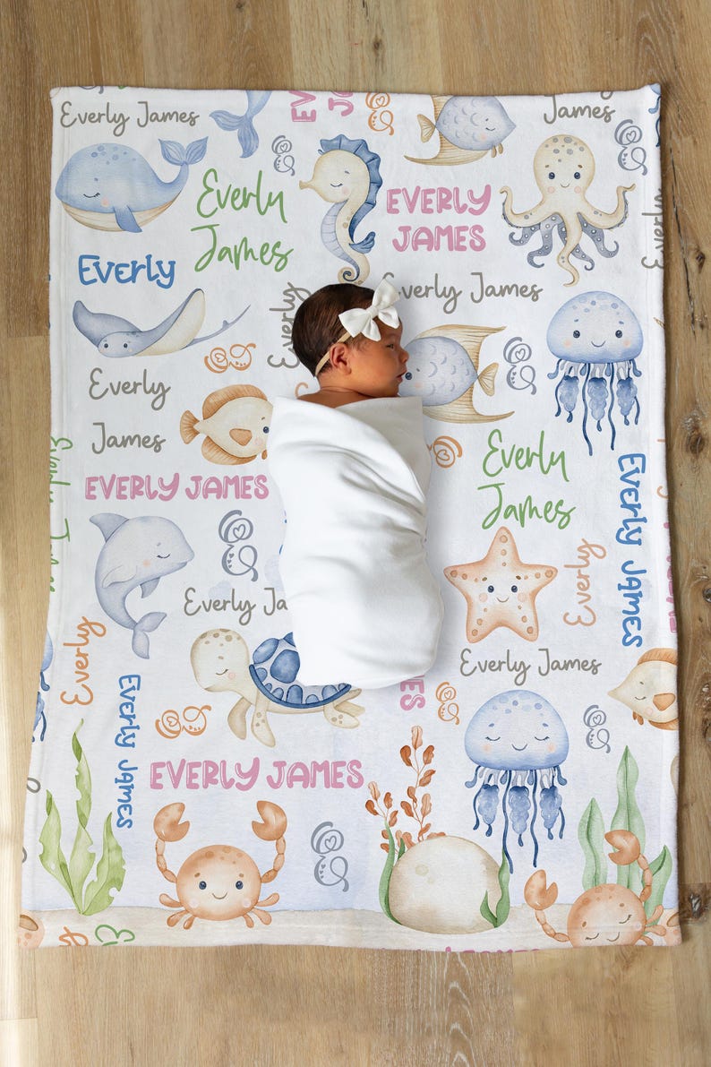May include: A white baby blanket with a sea life theme. The blanket features a variety of sea creatures, including a whale, a seahorse, an octopus, a jellyfish, a starfish, a turtle, and a crab. The blanket is decorated with the name "Everly James" repeated throughout.