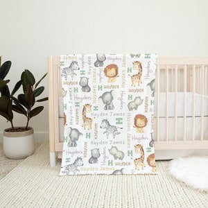 K&ouml;nnte beinhalten: Eine wei&szlig;e Babydecke mit einem sich wiederholenden Muster aus Safaritieren, darunter eine Giraffe, ein Zebra, ein L&ouml;we, ein Elefant und ein Nashorn. Die Tiere sind umgeben von dem Namen "Hayden James" in gr&uuml;ner und lila Schrift.
