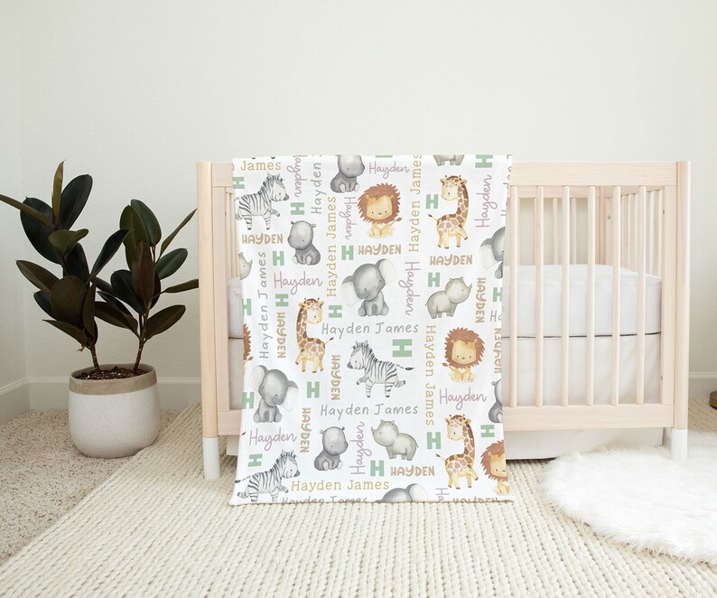 K&ouml;nnte beinhalten: Eine wei&szlig;e Babydecke mit einem sich wiederholenden Muster aus Safaritieren, darunter eine Giraffe, ein Zebra, ein L&ouml;we, ein Elefant und ein Nashorn. Die Tiere sind umgeben von dem Namen "Hayden James" in gr&uuml;ner und lila Schrift.