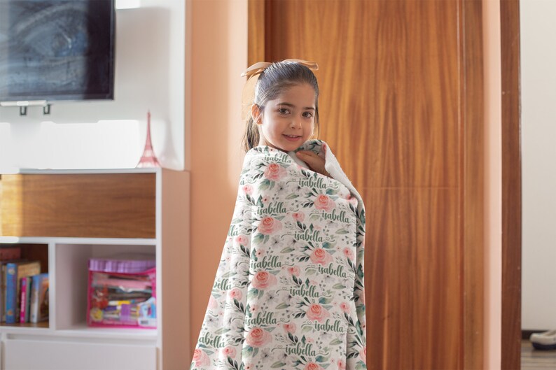 May include: A young girl is wrapped in a white blanket with a floral pattern and the name "Isabella" repeated throughout. The blanket has pink flowers and green leaves.