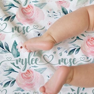 May include: A white baby blanket with a floral pattern featuring pink and green watercolour roses. The blanket has the names "Rylee Rose" written in green cursive with hearts.