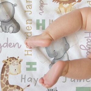 K&ouml;nnte beinhalten: Eine wei&szlig;e Babydecke mit einem hellgr&uuml;nen, braunen und grauen Tierdruck. Die Decke zeigt einen L&ouml;wen, eine Giraffe, ein Zebra, einen Elefanten und ein Flusspferd. Die Namen Hayden, James und Hay sind auf der Decke geschrieben.