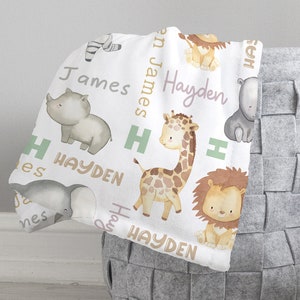 K&ouml;nnte beinhalten: Wei&szlig;e Babydecke mit einem sich wiederholenden Muster aus Tieren, darunter eine Giraffe, ein Nashorn, ein L&ouml;we, ein Elefant und ein Zebra. Die Decke tr&auml;gt die Namen Hayden und James in hellrosa und goldfarben.