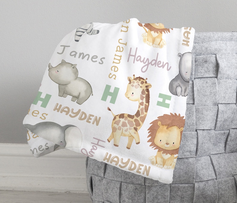 K&ouml;nnte beinhalten: Wei&szlig;e Babydecke mit einem sich wiederholenden Muster aus Tieren, darunter eine Giraffe, ein Nashorn, ein L&ouml;we, ein Elefant und ein Zebra. Die Decke tr&auml;gt die Namen Hayden und James in hellrosa und goldfarben.