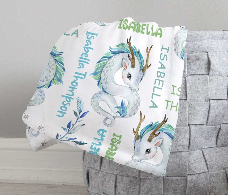 May include: White baby blanket with a repeating pattern of blue and green watercolor dragons. The blanket has the name "Isabella Thompson" written on it.