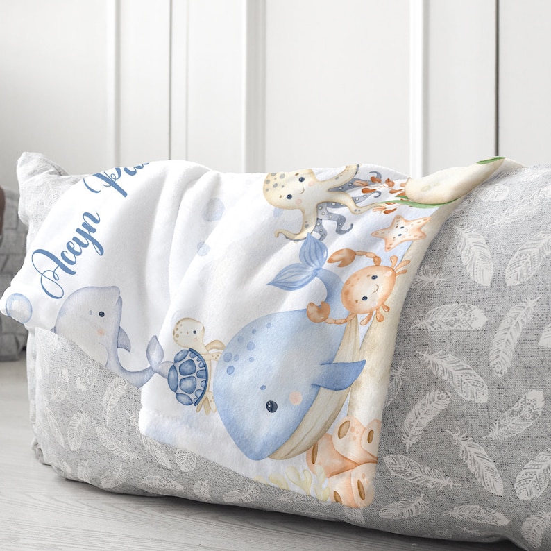 May include: A white baby blanket featuring a blue whale, octopus, turtle, crab and starfish design. The blanket is personalised with the name "Aeryn".