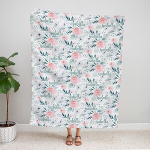 May include: A white fabric baby blanket with a repeating pattern of pink flowers, green leaves, and the name "isabella" written in cursive.