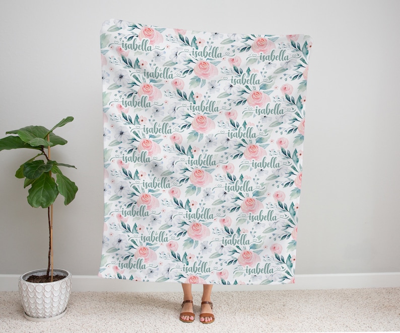 May include: A white fabric baby blanket with a repeating pattern of pink flowers, green leaves, and the name "isabella" written in cursive.