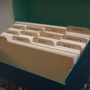 May include: A dark teal storage box containing white index cards with brown tabbed dividers. The tabs are labeled with black text: Spring, Summer, Fall, Winter, Monthly, To Do, Lists, Projects, Weekly, Week 1, and Week 2 for organization.