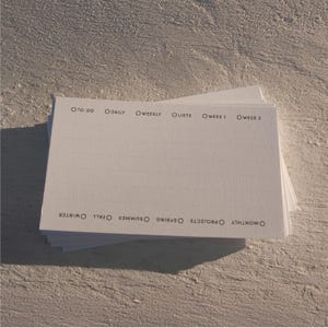 May include: A stack of rectangular white paper cards featuring black printed text and circular checkboxes. The top labels include TO DO, DAILY, WEEKLY, LISTS, WEEK 1, and WEEK 2. The bottom labels include MONTHLY, PROJECTS, SPRING, SUMMER, FALL, and WINTER.