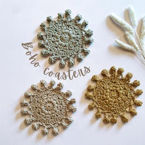Op de afbeelding: Drie handgemaakte gehaakte onderzetters in groen- en bruintinten. De onderzetters hebben een cirkelvormig ontwerp met een centraal patroon en een decoratieve rand. De woorden "Boho coasters" zijn cursief geschreven.