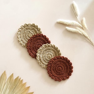 Op de afbeelding: Vier handgemaakte gehaakte onderzetters in beige en bruine tinten. De onderzetters hebben een ronde vorm met een geschulpte rand. Ze zijn diagonaal geplaatst op een lichtroze oppervlak, met gedroogde planten op de achtergrond.