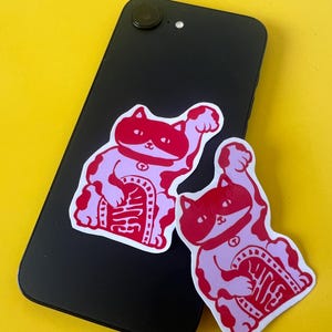 May include: Two red and pink lucky cat stickers on a black smartphone, against a yellow backdrop. The stickers show a traditional Japanese design with a raised paw and a panel with Japanese characters.