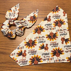 May include: A white fabric scrunchie and a white bandana with a brown and orange turkey print. The bandana has the text "Gobble Til You Wobble!" repeated on it.
