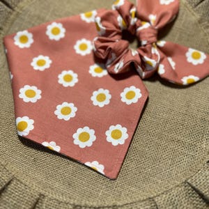 May include: A pink fabric scrunchie with a white daisy pattern. The scrunchie is tied in a bow and is sitting on a brown burlap surface.