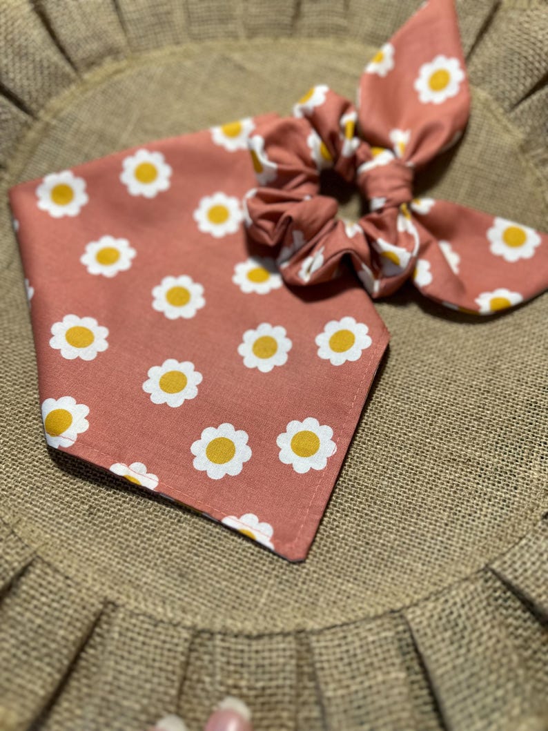 May include: A pink fabric scrunchie with a white daisy pattern. The scrunchie is tied in a bow and is sitting on a brown burlap surface.