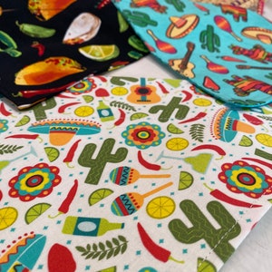 May include: Four fabric squares with different Mexican-themed patterns. The patterns include a black square with yellow and red tacos, a turquoise square with cacti and maracas, a white square with cacti, maracas, and limes, and a white square with a guitar, limes, and chili peppers.