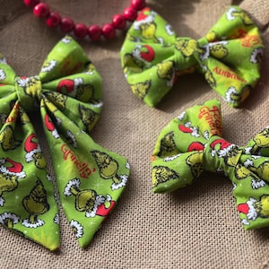 May include: Three green fabric hair bows with a Grinch-themed pattern. The bows feature the Grinch character in a Santa hat, with red and white accents. A red beaded necklace is also in the image.