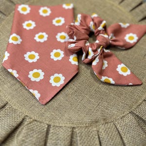 May include: A set of two pink and white floral patterned accessories. A bandana with white daisies and yellow centers on a pink background and a matching scrunchie.