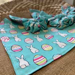May include: A turquoise blue bandana with a white bunny rabbit and colorful Easter egg pattern. The bandana is folded into a bow shape and is perfect for a pet.