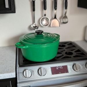 Pode incluir: Uma cena de cozinha em miniatura com uma panela verde vibrante sobre um fogão prateado. Acima, um suporte contém pequenos utensílios de cozinha. O visor digital do fogão mostra a hora 12:45. A panela tem cerca de 15 cm de diâmetro.