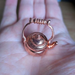 Puede incluir: Una pequeña tetera de color cobre con un cuerpo redondeado, un pequeño pico y un asa en forma de lazo. El asa presenta un agarre texturizado. La tetera es un pequeño objeto decorativo, posiblemente un amuleto o un objeto de colección.