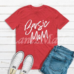 May include: Red t-shirt with white text that says "Basic Mom". The shirt is laying on a wooden surface with a pair of white sneakers and blue jeans.
