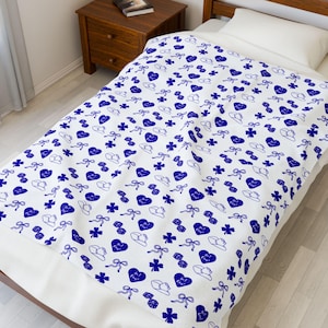 May include: A white blanket with a repeating pattern of blue hearts, bows, hats, and clovers. The blanket is spread over a bed with a white pillow. The bed is made of wood.