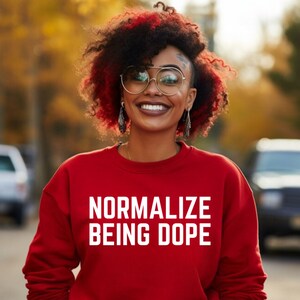 Puede incluir: Una sudadera roja con el texto "Normalize Being Dope" en letras blancas. La modelo lleva la sudadera y tiene el pelo rojo y negro.