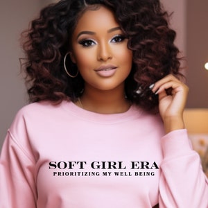 Puede incluir: Una sudadera rosa con el texto "SOFT GIRL ERA PRIORITIZING MY WELL BEING" impreso en la parte delantera.