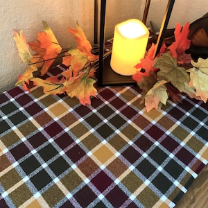 May include: A plaid table runner featuring a pattern of dark green, burgundy, mustard yellow, and white squares. A black metal lantern with a glowing LED candle sits on the runner, surrounded by artificial orange and yellow maple leaves.