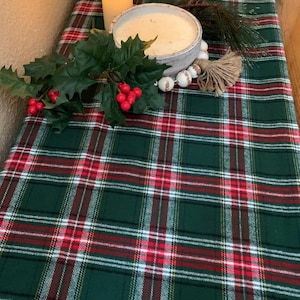 May include: A green, red, and white plaid table runner with a white candle in a bowl and a sprig of greenery with red berries.