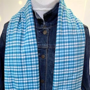 May include: A light blue and white gingham patterned scarf draped over a dark blue denim jacket and a white ribbed turtleneck sweater. The scarf features a consistent checkered design in varying shades of blue and white, worn around the neck.