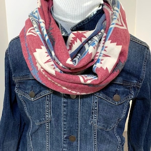 May include: A red, white, and blue patterned infinity scarf. The scarf is wrapped around a white turtleneck and a denim jacket. The pattern features geometric shapes and designs.