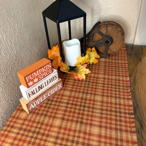 May include: A black metal lantern with a white pillar candle, surrounded by orange and yellow faux leaves. Next to it are three stacked wooden blocks labeled PUMPKIN SPICE, FALLING LEAVES, and APPLE CIDER, resting on a brown and orange plaid runner.