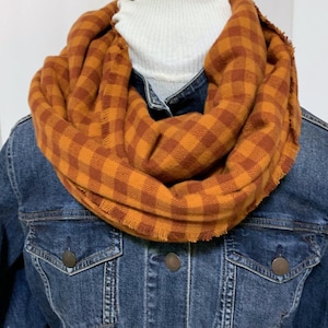 May include: An orange and brown plaid infinity scarf with a fringed edge. The scarf is draped over a white turtleneck and a denim jacket. The scarf's pattern is a classic buffalo check, perfect for fall and winter.