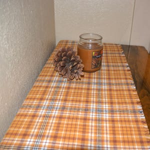 May include: A brown and white plaid table runner with frayed edges. A pine cone and a glass jar with a candle inside are on the runner.