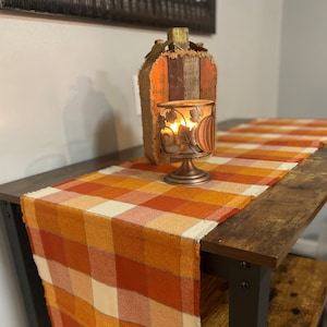 May include: A brown wooden table with a plaid table runner in shades of orange, cream, and brown. A metal candle holder with a pumpkin design sits on the table runner. The candle holder has a lit candle inside.