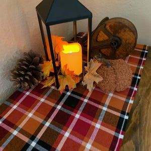 May include: A black metal lantern containing an illuminated orange LED candle, surrounded by artificial autumn leaves, a pinecone, a textured brown fabric pumpkin, and a vintage wooden pulley, all resting on a red, orange, and black plaid table runner.