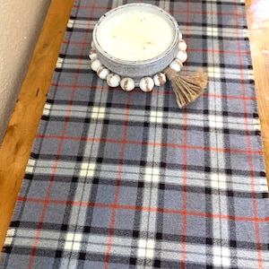 May include: A gray and white plaid table runner with orange accents. The runner has a frayed edge and is decorated with a white ceramic bowl and a wooden bead garland.