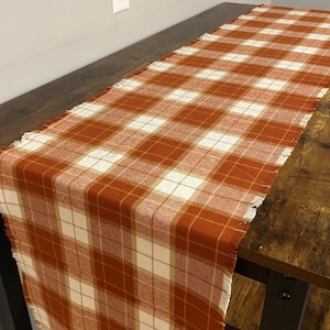 May include: A rust orange and white plaid table runner with frayed edges. The runner is made of a soft, woven fabric and is perfect for adding a touch of rustic charm to any table setting.