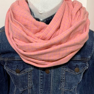May include: A soft pink infinity scarf featuring a subtle grid pattern in thin purple and orange lines. The scarf is draped in multiple layers around the neck, resting over a dark blue denim jacket with visible metal buttons and a white ribbed collar.