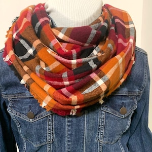 May include: A warm, cozy scarf in a plaid pattern with shades of orange, red, black, and white. The scarf is made of a soft, comfortable fabric and is perfect for fall and winter.