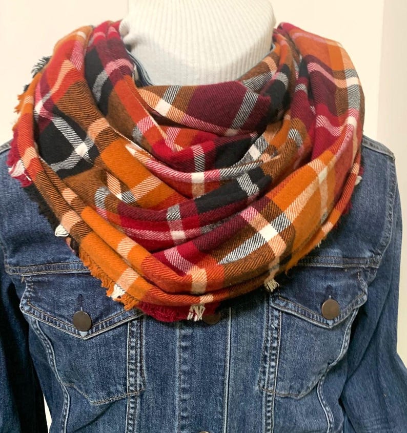 May include: A warm, cozy scarf in a plaid pattern with shades of orange, red, black, and white. The scarf is made of a soft, comfortable fabric and is perfect for fall and winter.