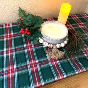 May include: A green and red plaid table runner with a white candle, a white ceramic bowl with a white beaded garland, and a sprig of holly with red berries.