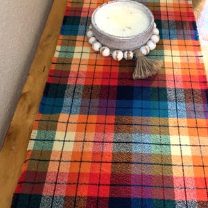May include: A plaid table runner with a frayed edge in shades of orange, red, blue, green, and cream. A white candle in a ceramic bowl sits on top of the runner, surrounded by a beaded garland.