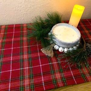 May include: A white ceramic candle holder with a white candle inside, surrounded by a pine garland and white beads. The candle holder is sitting on a red and white plaid table runner.