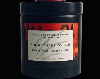 Könnte beinhalten: Ein schwarzes Kerzengefäß mit einem roten Wachstropfen-Design. Das Etikett lautet "MAGIC BOX CANDLE COMPANY - MADE IN ARIZONA - A NIGHTMARE ON ELM - FIRESIDE EMBERS + SUEDE + VETIVER - 9OZ | COCONUT SOY CANDLE".