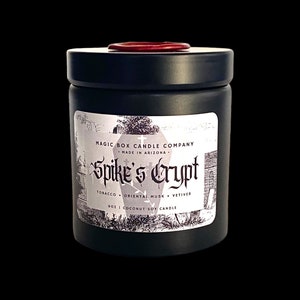 May include: Black candle jar with a red lid. The label reads "Magic Box Candle Company", "Made in Arizona", "Spike's Crypt", "Tobacco + Oriental Musk + Vetiver", and "9 oz | Coconut Soy Candle".