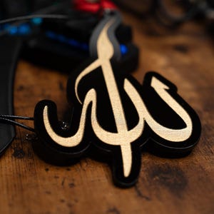 May include: A black and gold decorative ornament shaped like the Arabic word for God, "Allah". The ornament is hanging from a black cord.